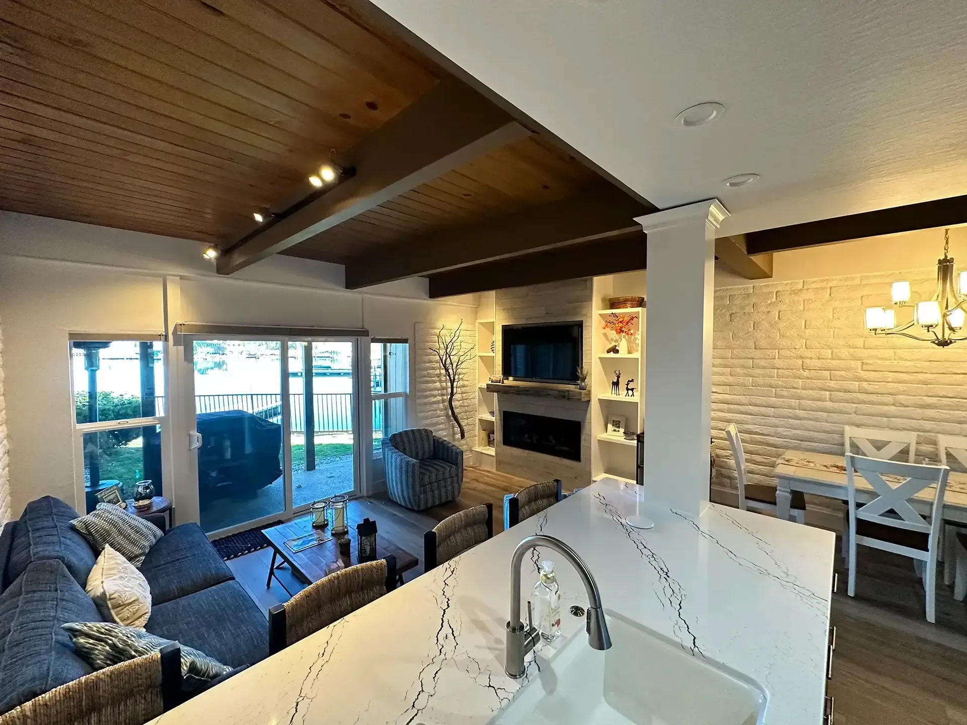 An open-concept living area with a kitchen island, blue sofa, fireplace, and dining space under wood-paneled ceilings.
