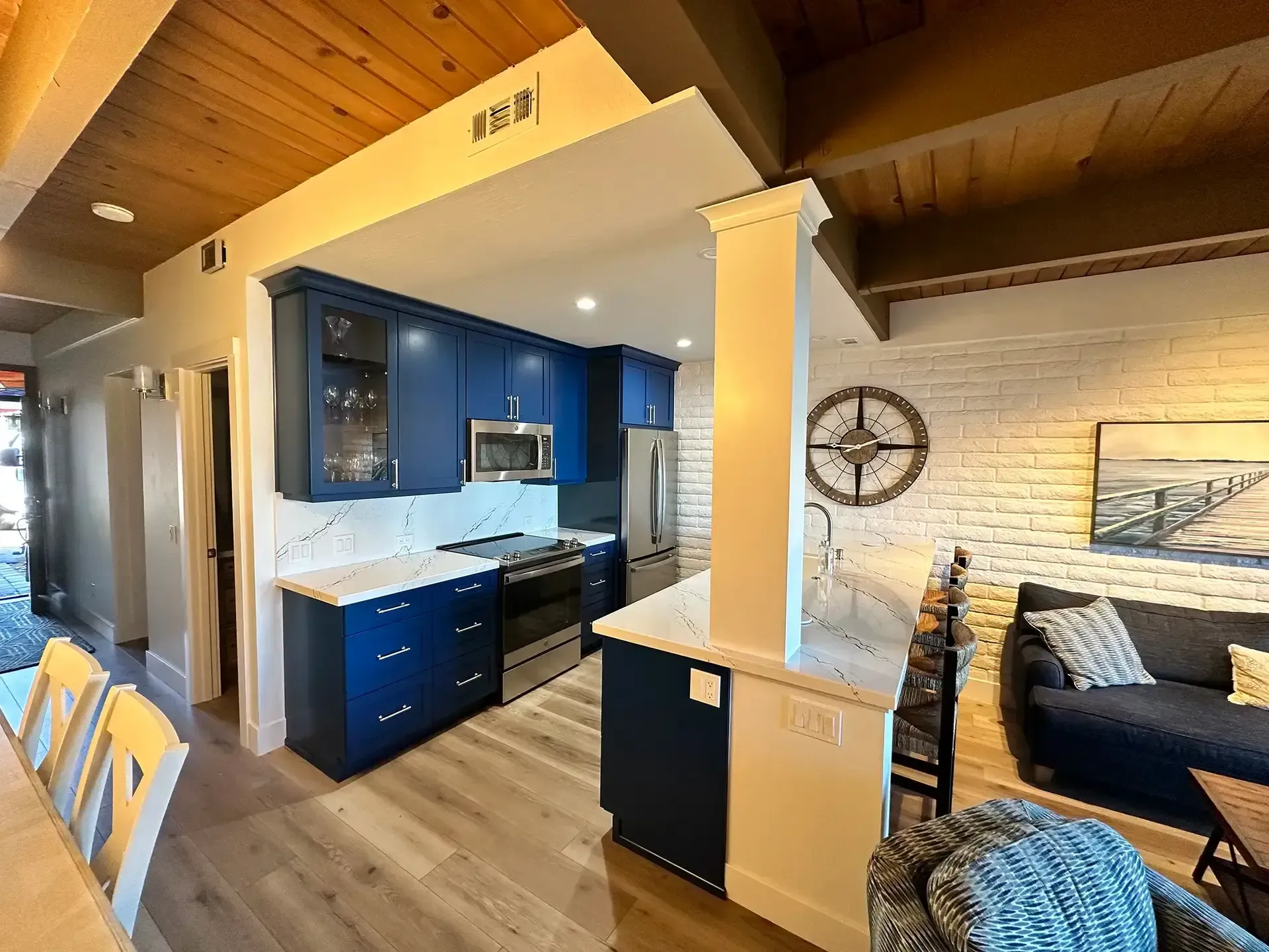 A modern kitchen with blue cabinets, white countertops, and wood ceilings, next to a living room with a white brick wall.