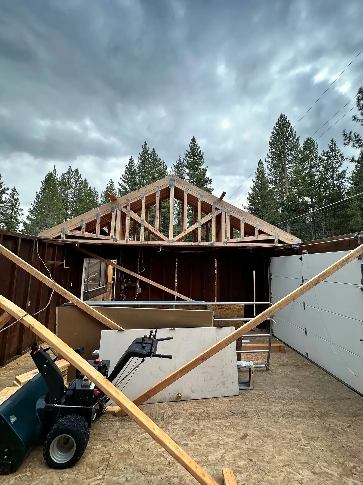 Construction in progress on a wooden roof frame with an open gable above a partially enclosed space with a snowblower.