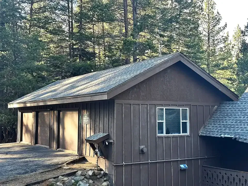 A brown wooden garage with a grey shingled roof, surrounded by tall pine trees.