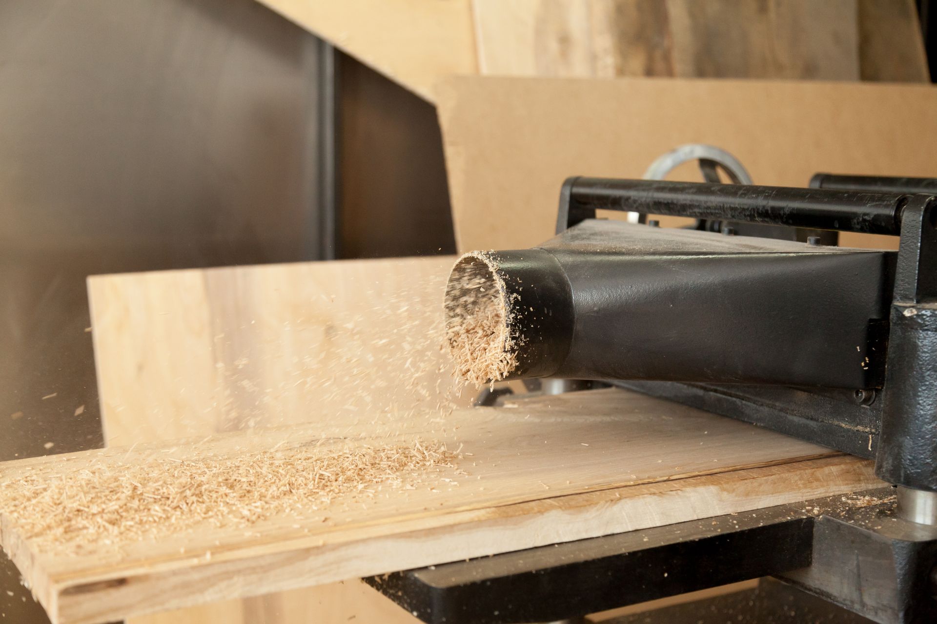 Wood shavings fly from the black dust collection port of a wood planer as it cuts into a piece of wooden board.