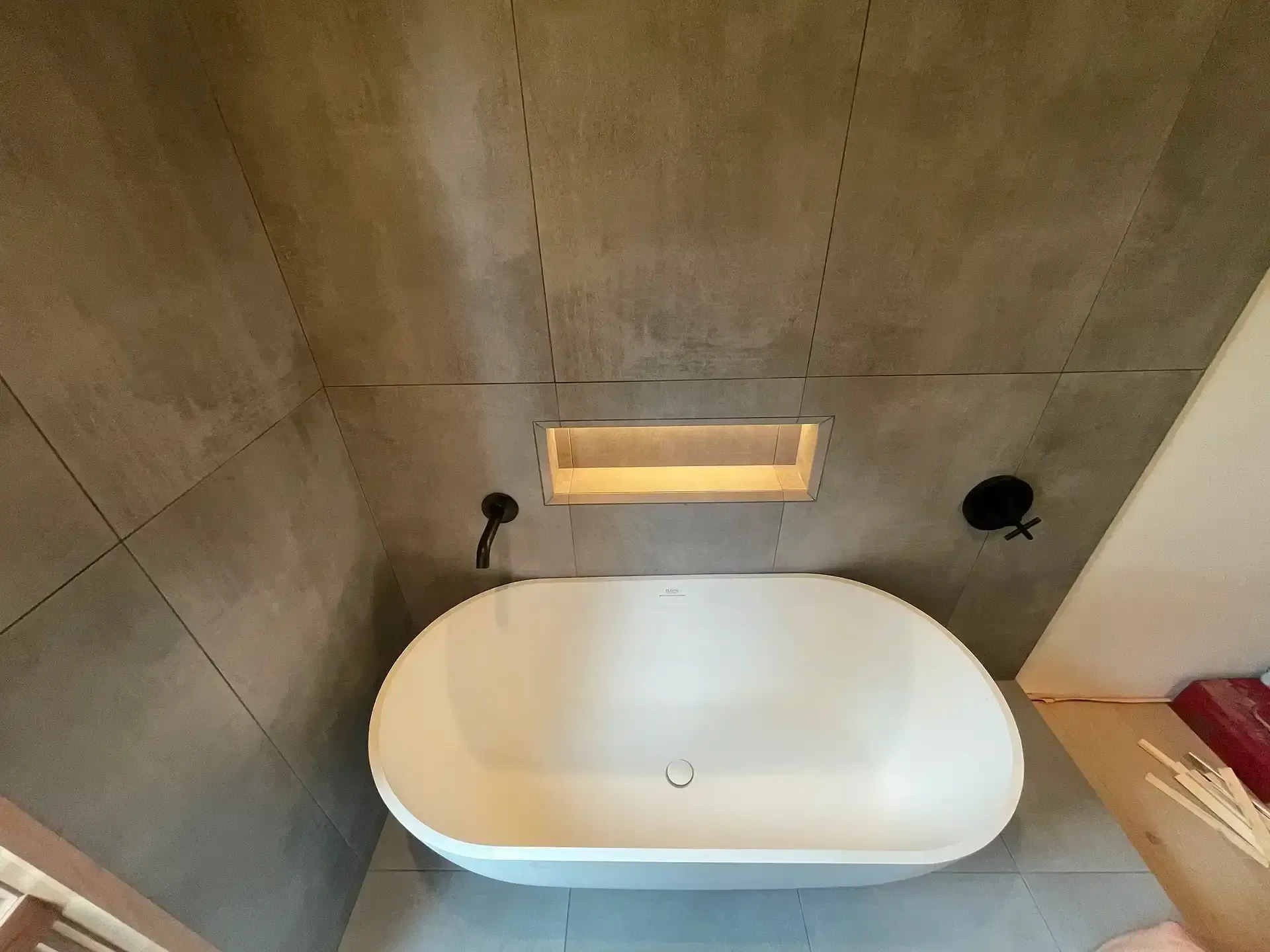 A white oval freestanding bathtub centered against a gray tiled wall with a recessed, illuminated storage niche.