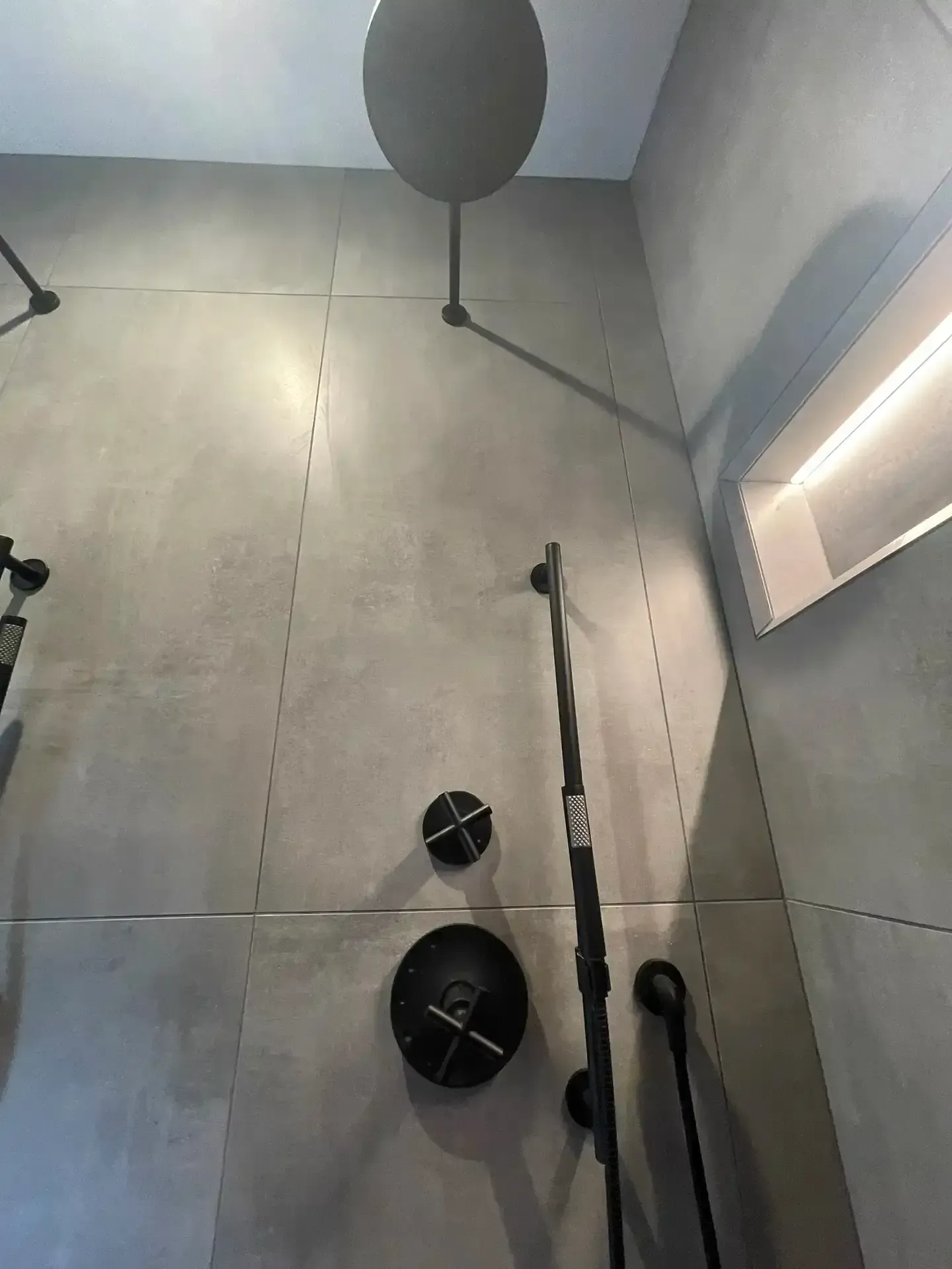 A modern shower with matte black fixtures, a round rainfall showerhead, a handheld sprayer, and gray concrete-style tiles.