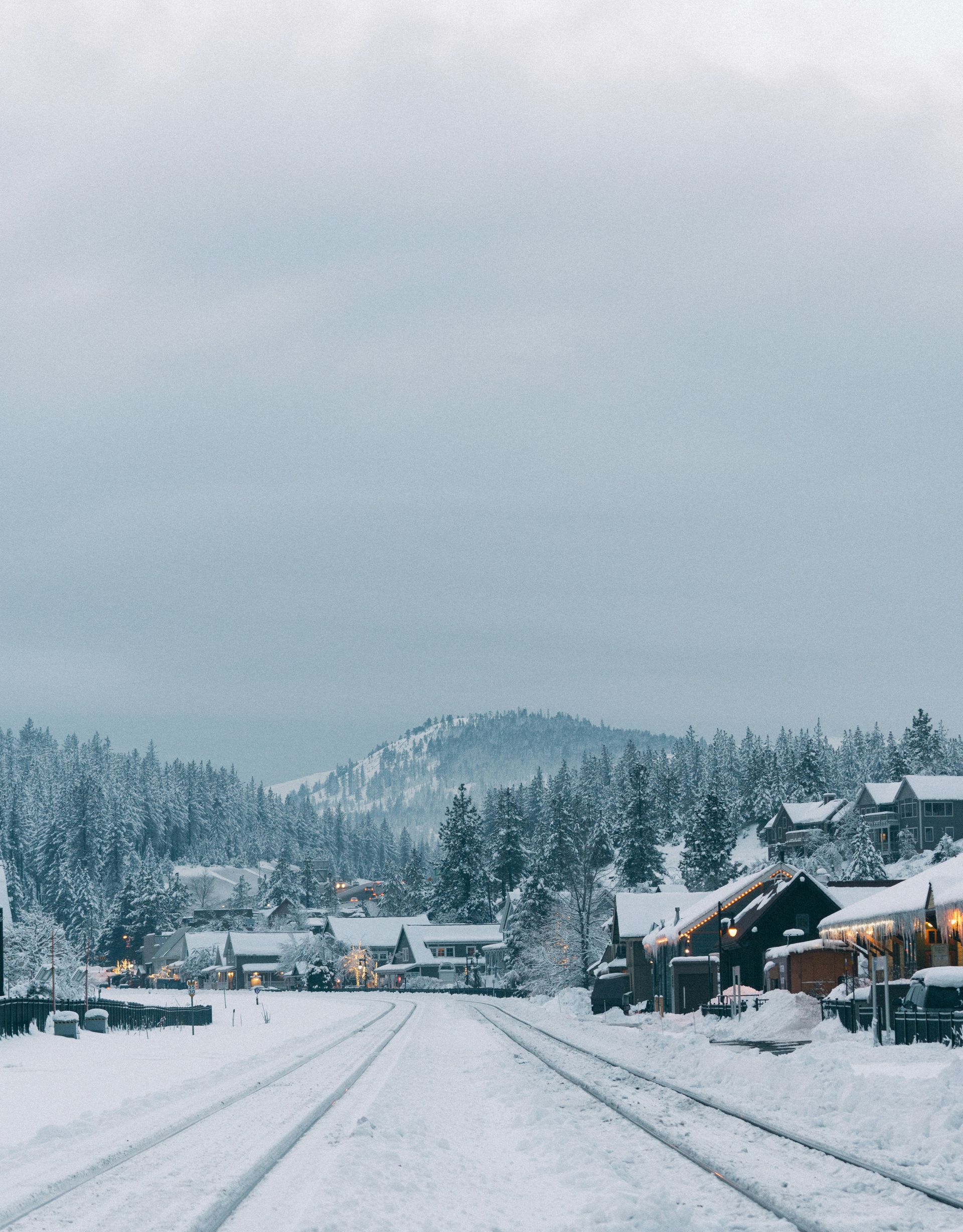 Snow-covered road in a mountain town with houses, trees, and cloudy sky