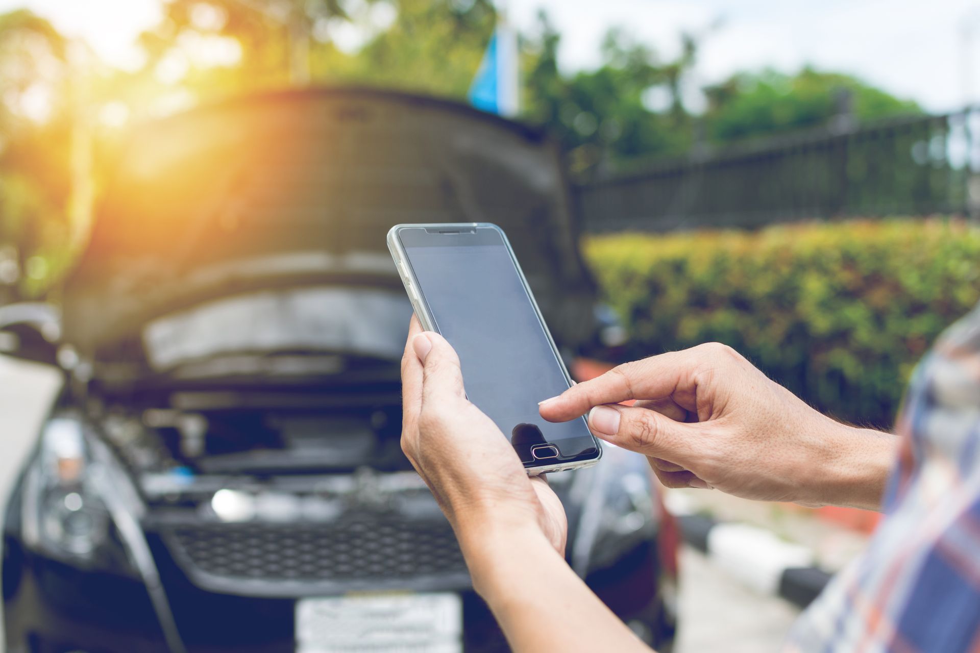 Person using a smartphone in front of an open car hood, checking vehicle issue outdoors.