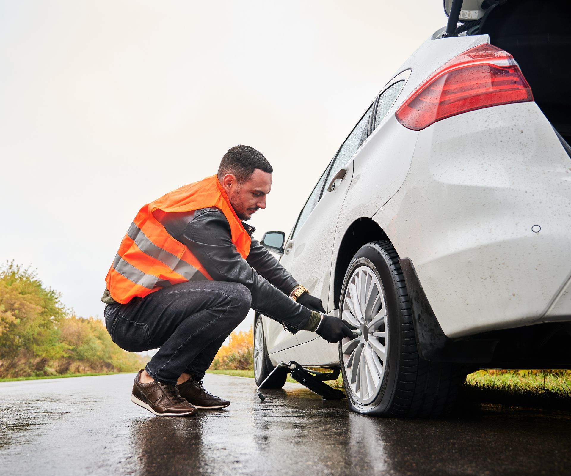 Man in an orange safety vest jacks up a white car to change a tire during 24/7 roadside assistance.