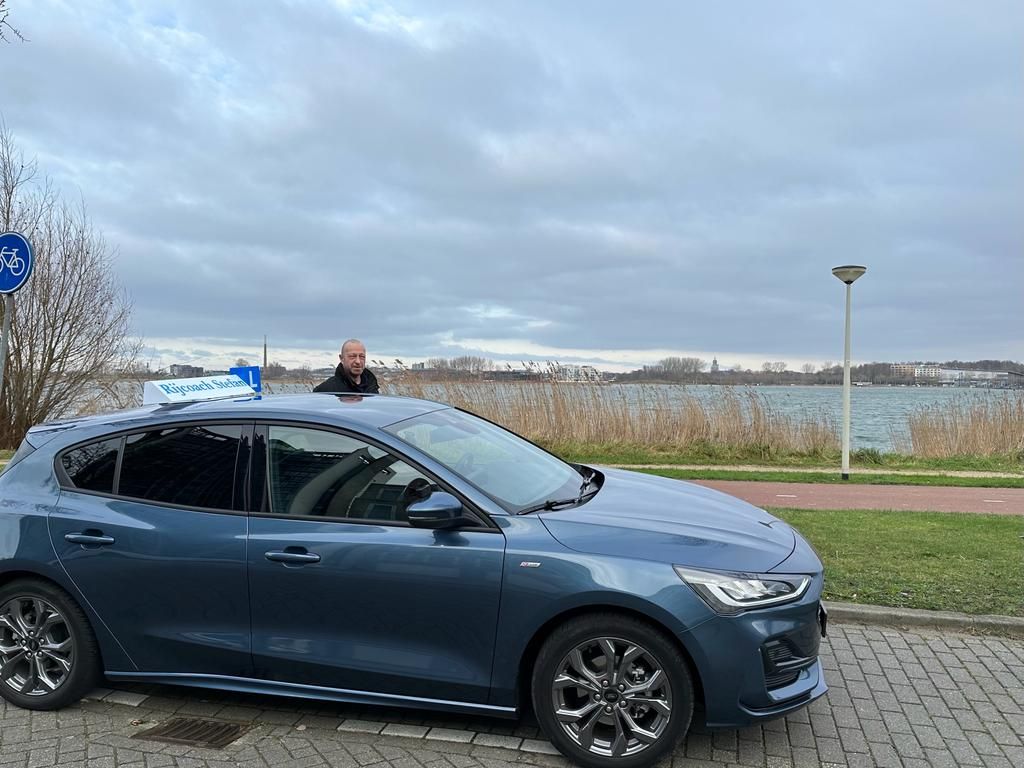 Een blauwe auto staat geparkeerd bij een waterpartij; een persoon staat achter de auto, onder een bewolkte hemel.