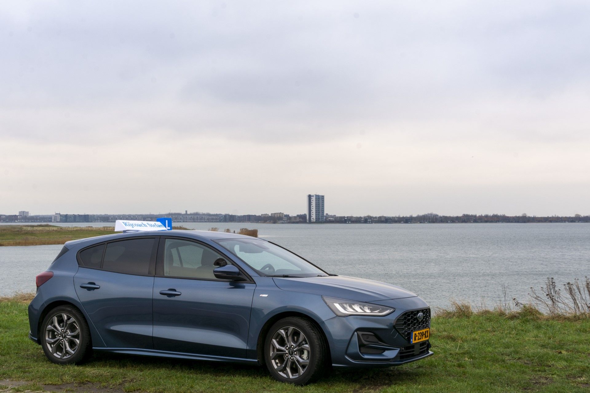 Een blauwe Ford Focus staat geparkeerd aan het water, met een bord van een rijschool op het dak. 