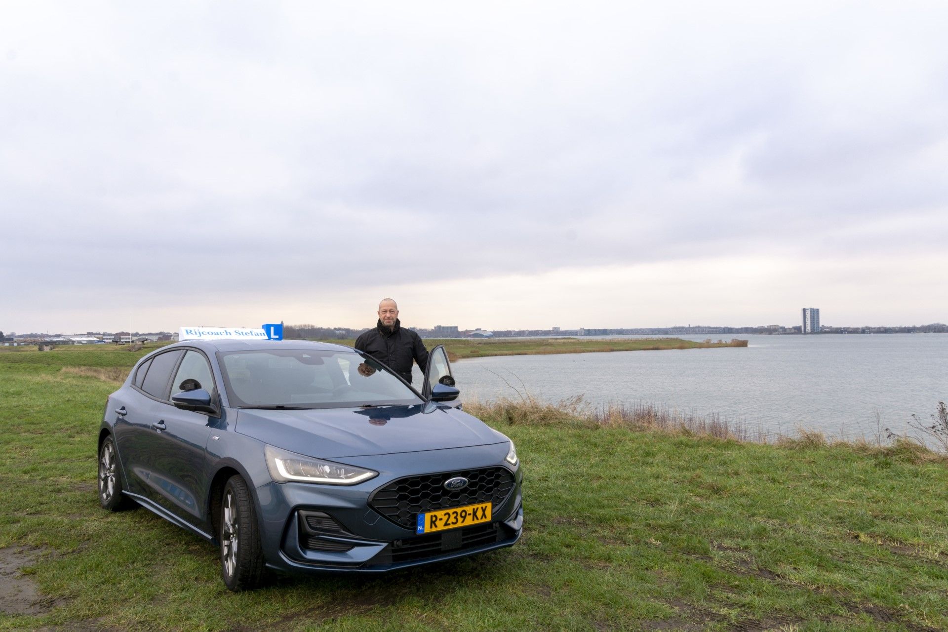 Een man staat bij een donkerblauwe Ford met open deur, vlakbij het water, onder een bewolkte hemel.