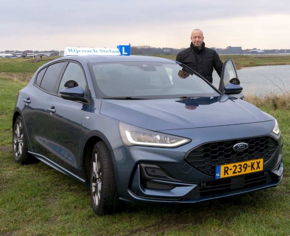 Een man staat naast een blauwe Ford Focus, met een open deur. Op het dak staat een bord van een rijschool.