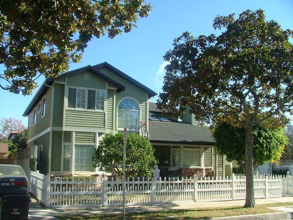 Green two-story house with white picket fence and trees on a sunny day.