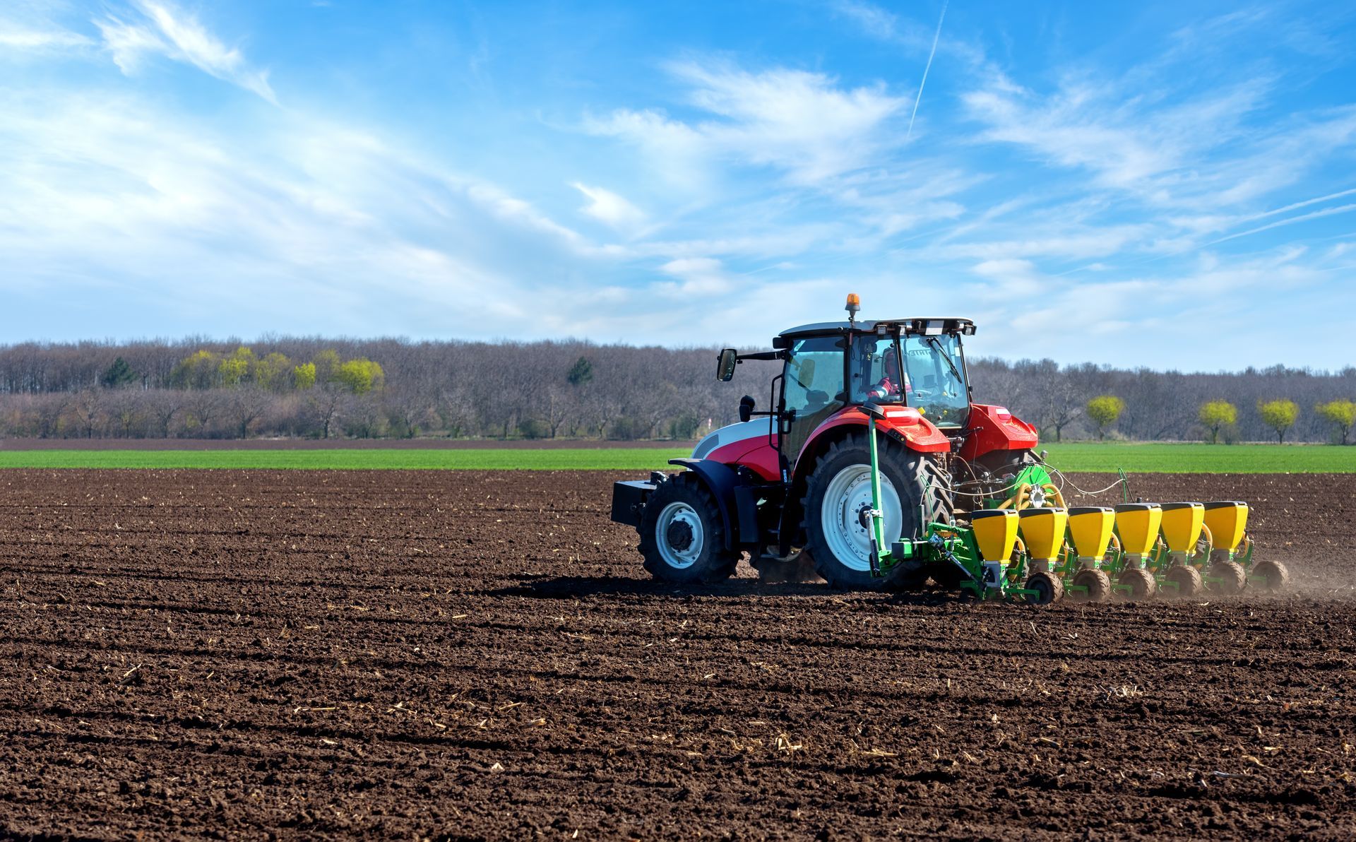 Tractor plowing a field with a seed drill under a bright blue sky.