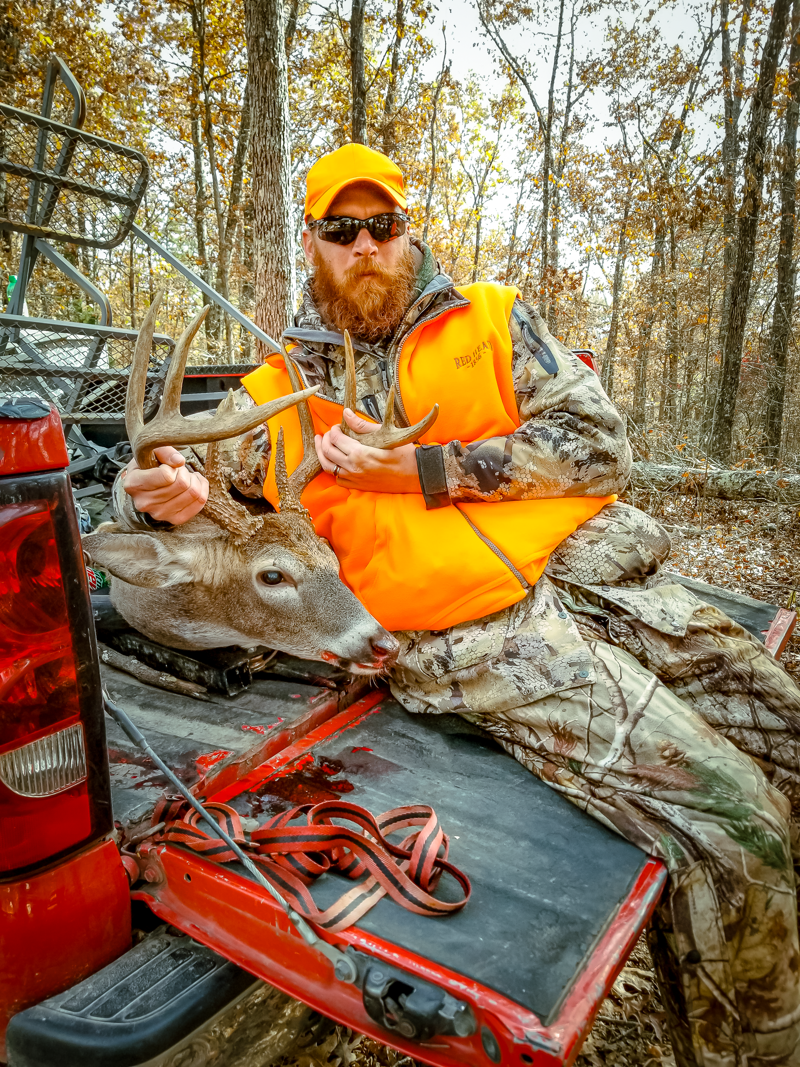 Man in orange hunting vest sits beside a deer in a truck bed. He holds the deer's antlers. Fall foliage in background.