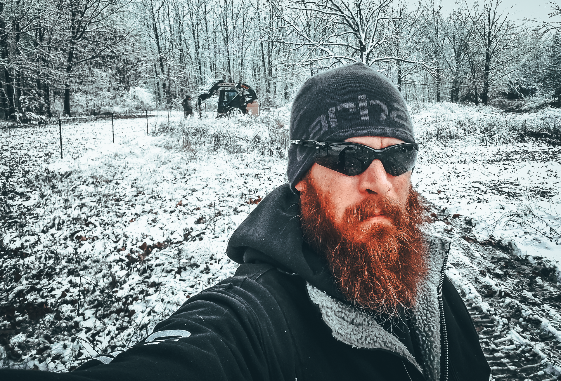 Man with red beard, sunglasses, and beanie in a snowy field, taking a selfie.