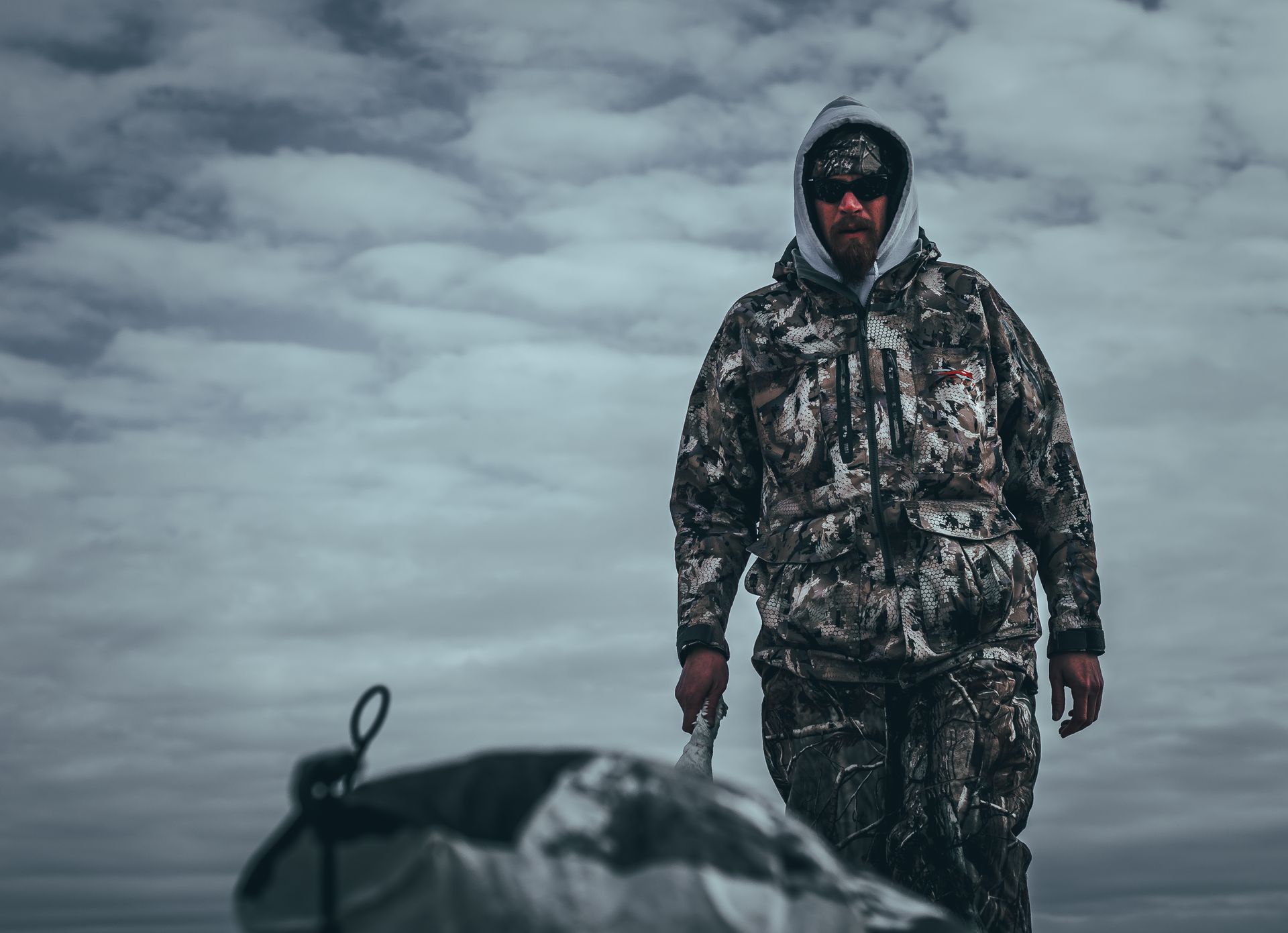 Person in camouflage jacket and pants, hooded, holds something, possibly a sled, on an overcast day.