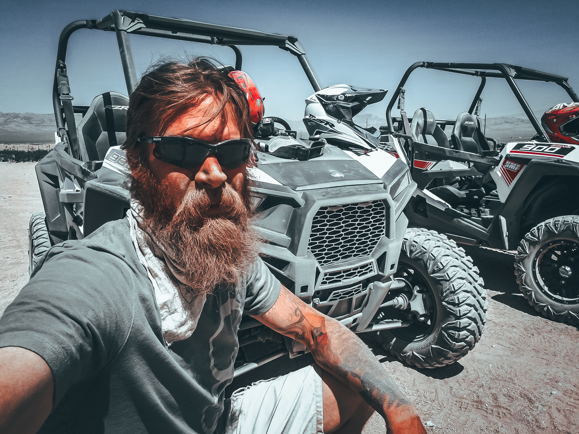 Man with beard and sunglasses, sits near two UTVs in a desert setting, taking a selfie.