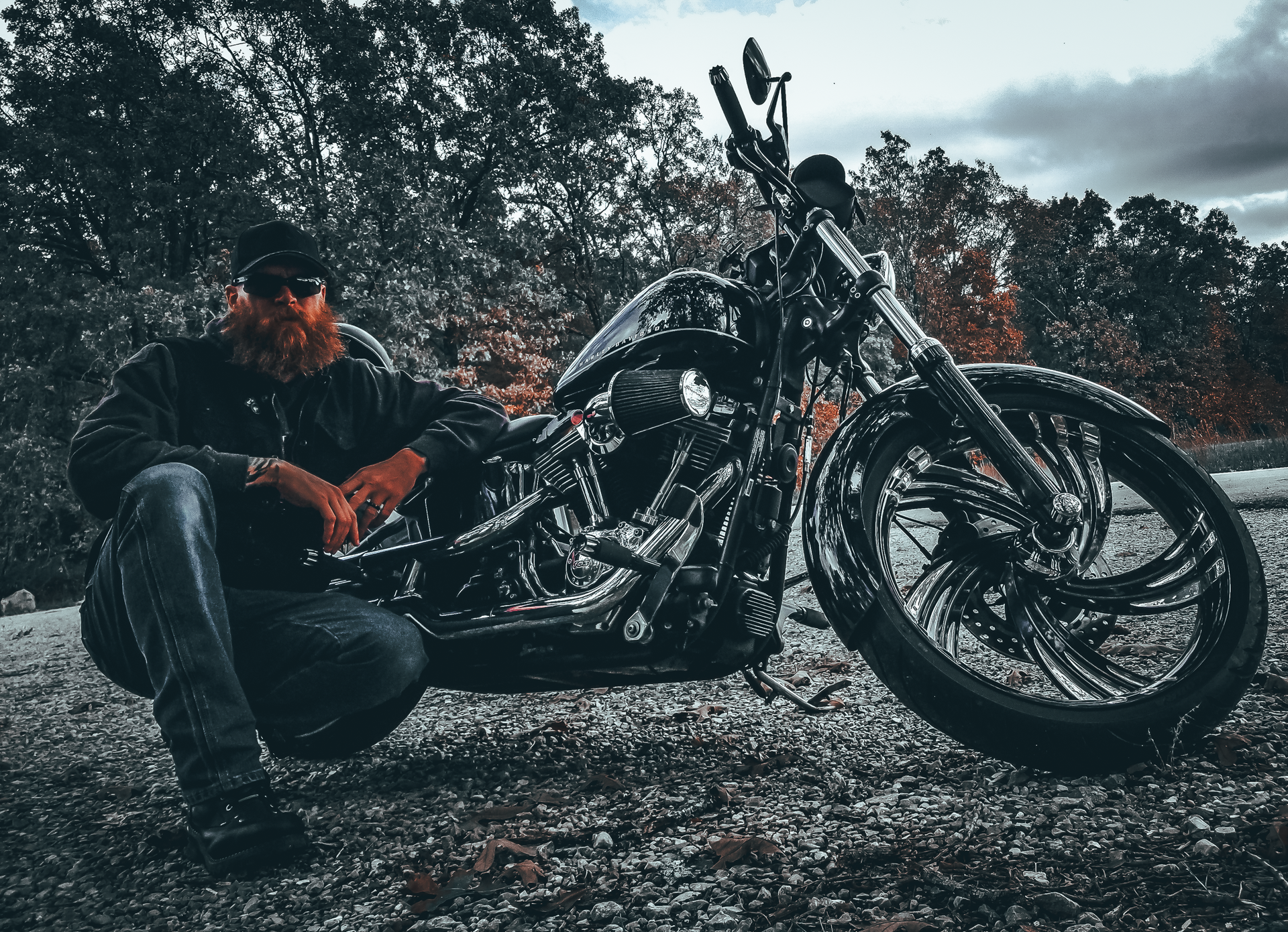 Man with large beard squats beside custom motorcycle outdoors.