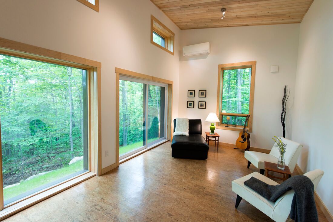 Salon lumineux avec de grandes fenêtres donnant sur les arbres ; sièges, guitare et œuvres d'art.