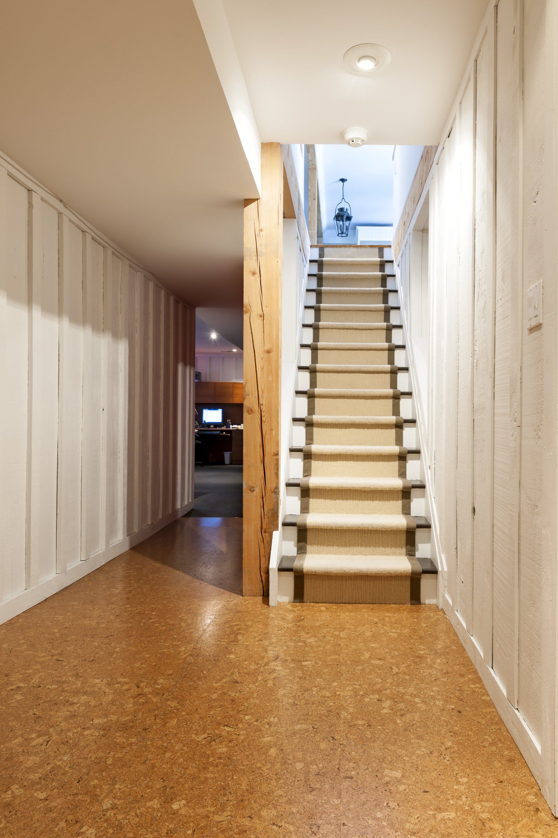 Couloir avec escalier et lambris clair, sol en liège. Un poteau en bois naturel se trouve à droite.