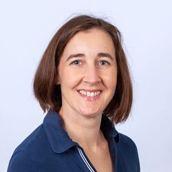 Eine Frau mit kurzen braunen Haaren lächelt vor weißem Hintergrund. Sie trägt ein dunkelblaues Poloshirt.