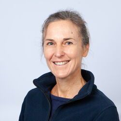 Frau lächelt, trägt eine blaue Reißverschlussjacke. Graues Haar zurückgebunden, hellblauer Hintergrund.