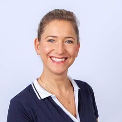 Eine hellhäutige Frau lächelt vor hellblauem Hintergrund. Sie trägt ein dunkelblaues Hemd mit weißem Kragen.