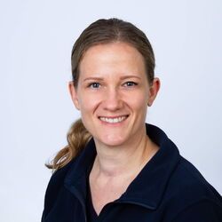 Frau mit hellbraunem, zurückgebundenem Haar, lächelnd, trägt eine dunkelblaue Jacke.