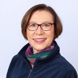 Frau mit Brille, lächelnd, trägt eine blaue Jacke und einen bunten Schal vor weißem Hintergrund.