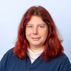 Frau mit roten Haaren, die ein blaues Hemd und einen weißen Rollkragenpullover trägt, lächelt.