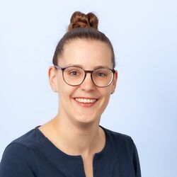Frau mit Brille und Dutt lächelt in die Kamera, trägt ein marineblaues Oberteil vor hellblauem Hintergrund.