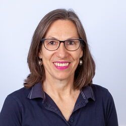 Frau mit Brille und rosa Lippenstift lächelt in die Kamera; sie trägt ein dunkelblaues Hemd.
