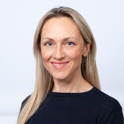 Frau mit langen blonden Haaren, die in die Kamera lächelt, trägt ein dunkelblaues Hemd vor weißem Hintergrund.