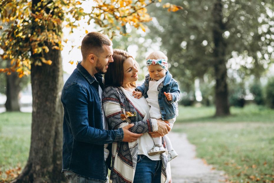 Family of three posing outdoors: parents holding baby, smiling in park setting with fall foliage.
