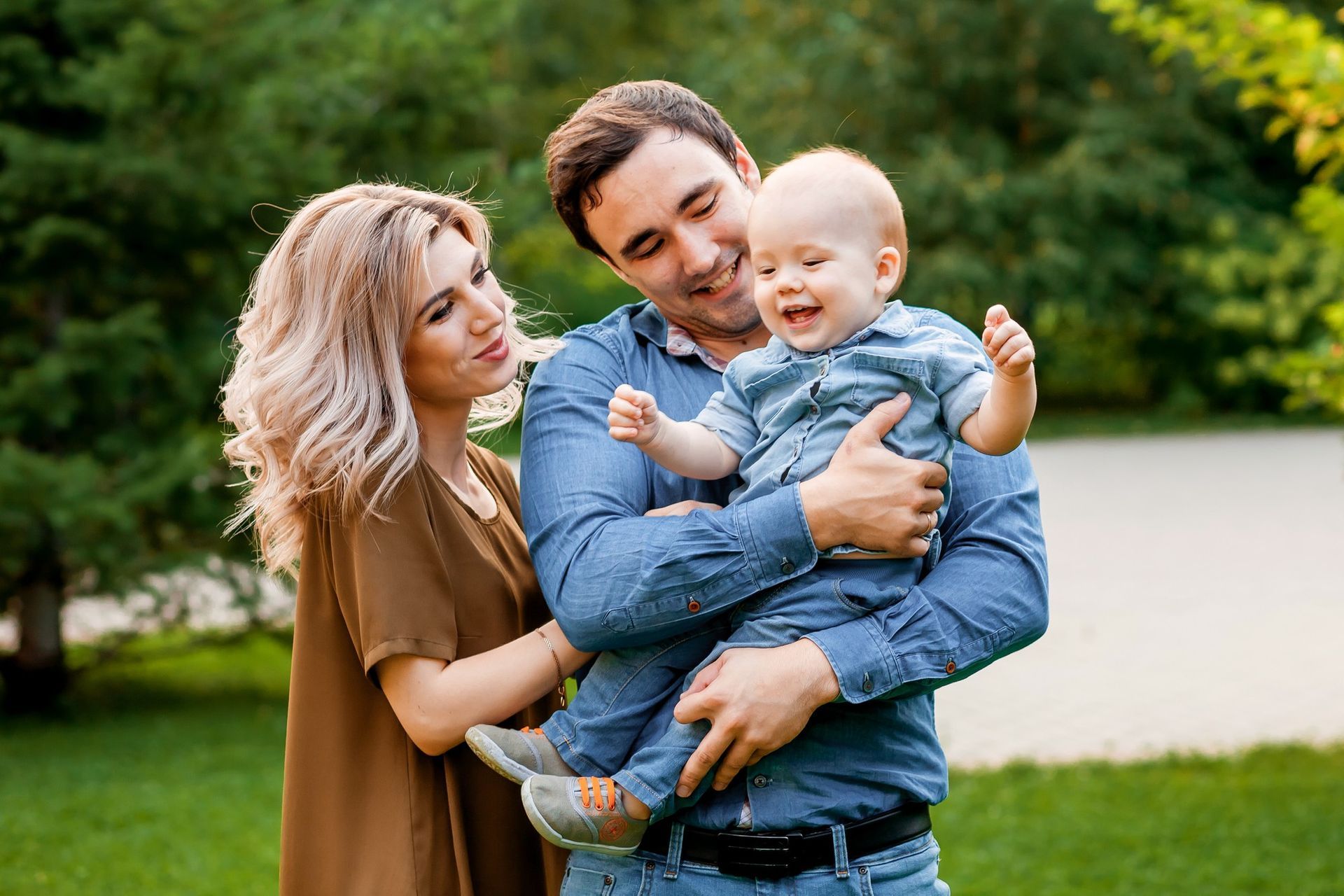 Parents holding a smiling baby outdoors, with green grass and trees in the background.