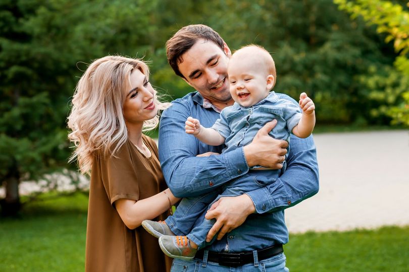 Parents holding a smiling baby outdoors, with green grass and trees in the background.