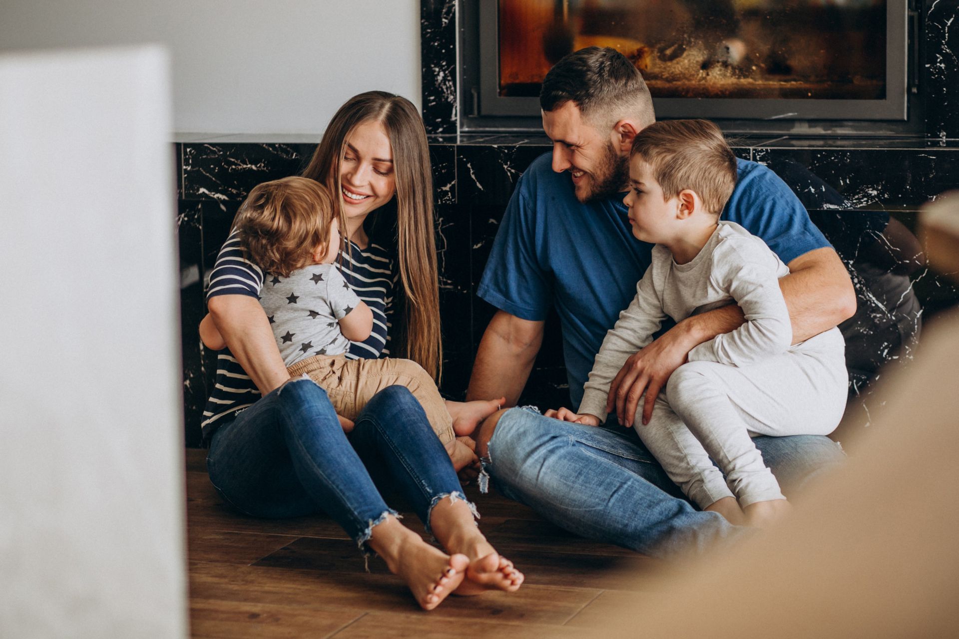 Family of four sitting on floor in front of fireplace, smiling and embracing each other.