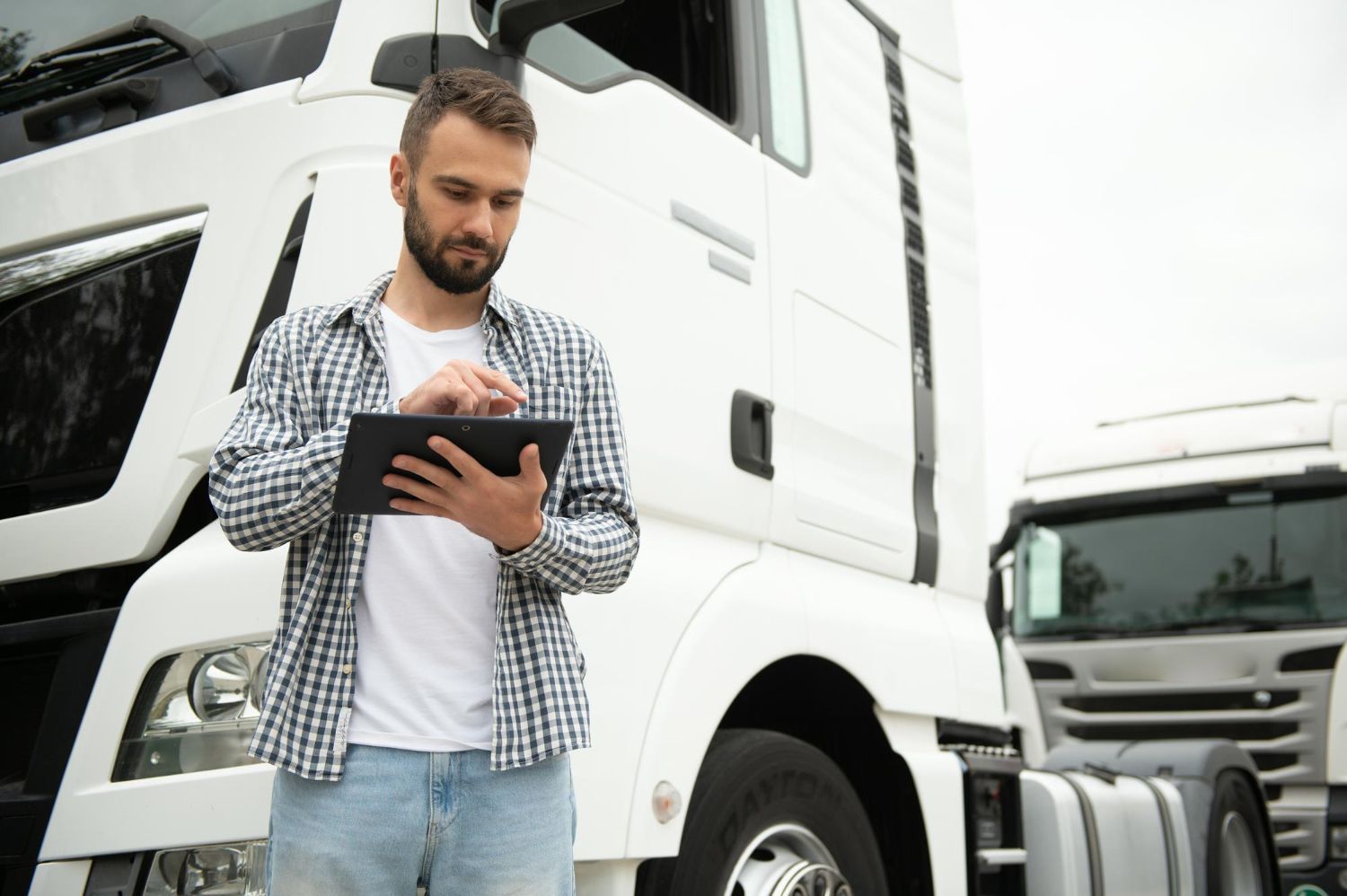 Man with tablet stands near white semi-trucks, likely checking or recording information.