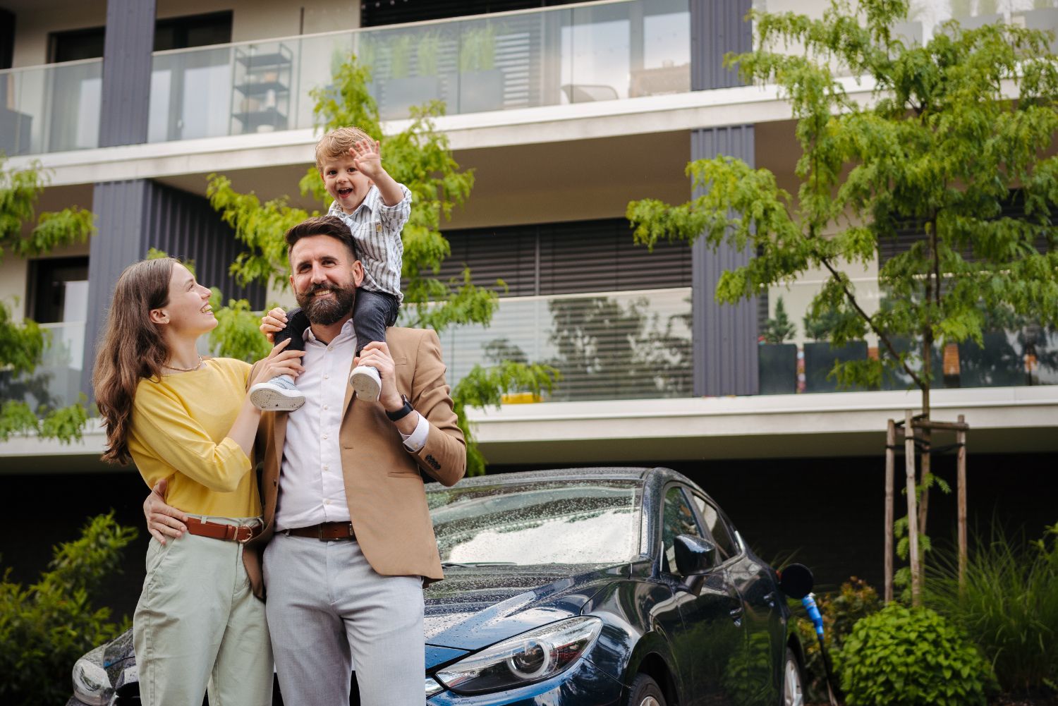 Family, with child on father’s shoulders, stands by a blue electric car in front of an apartment building.