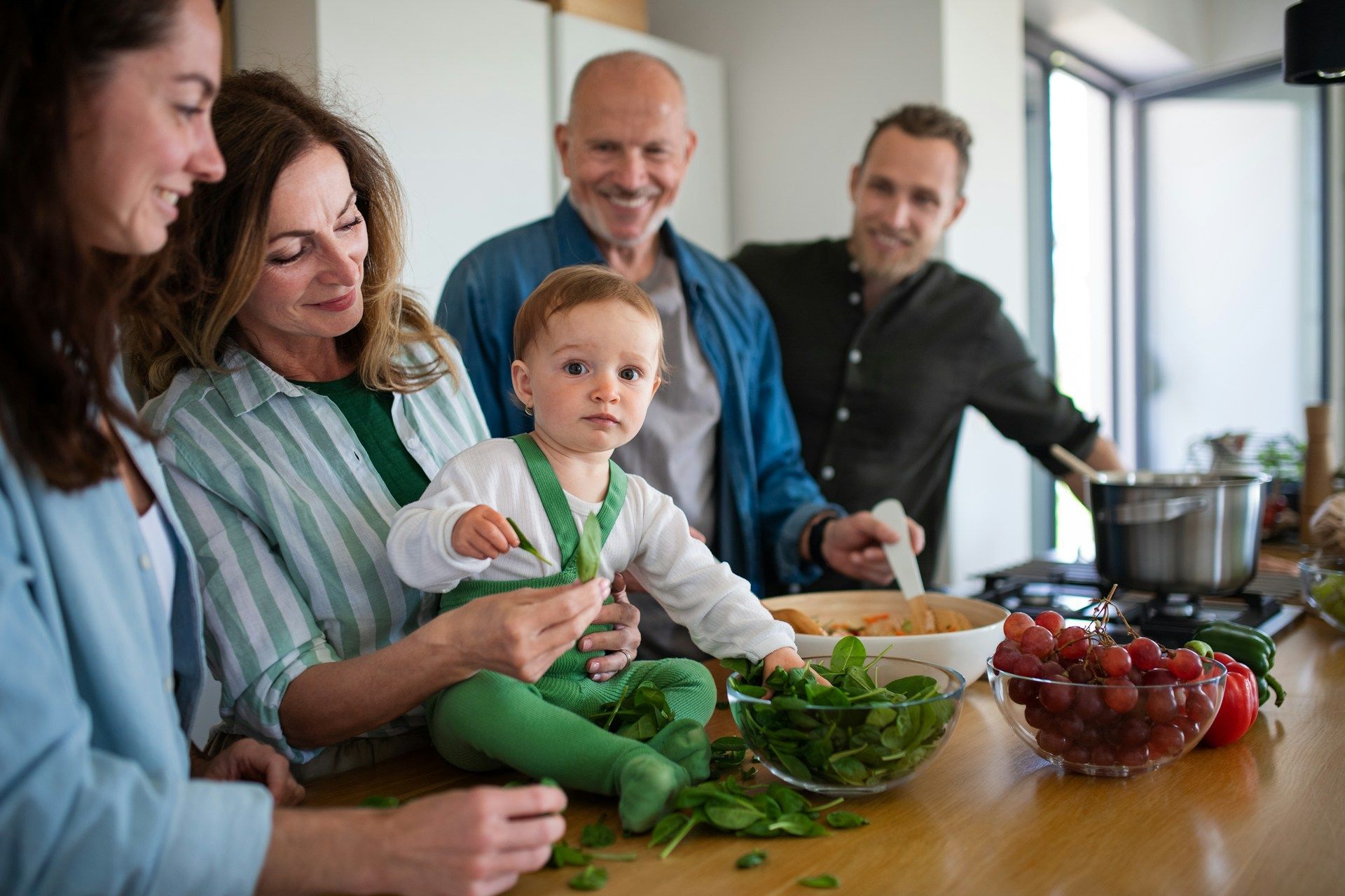 Family in kitchen: baby held by woman, others cooking near vegetables.