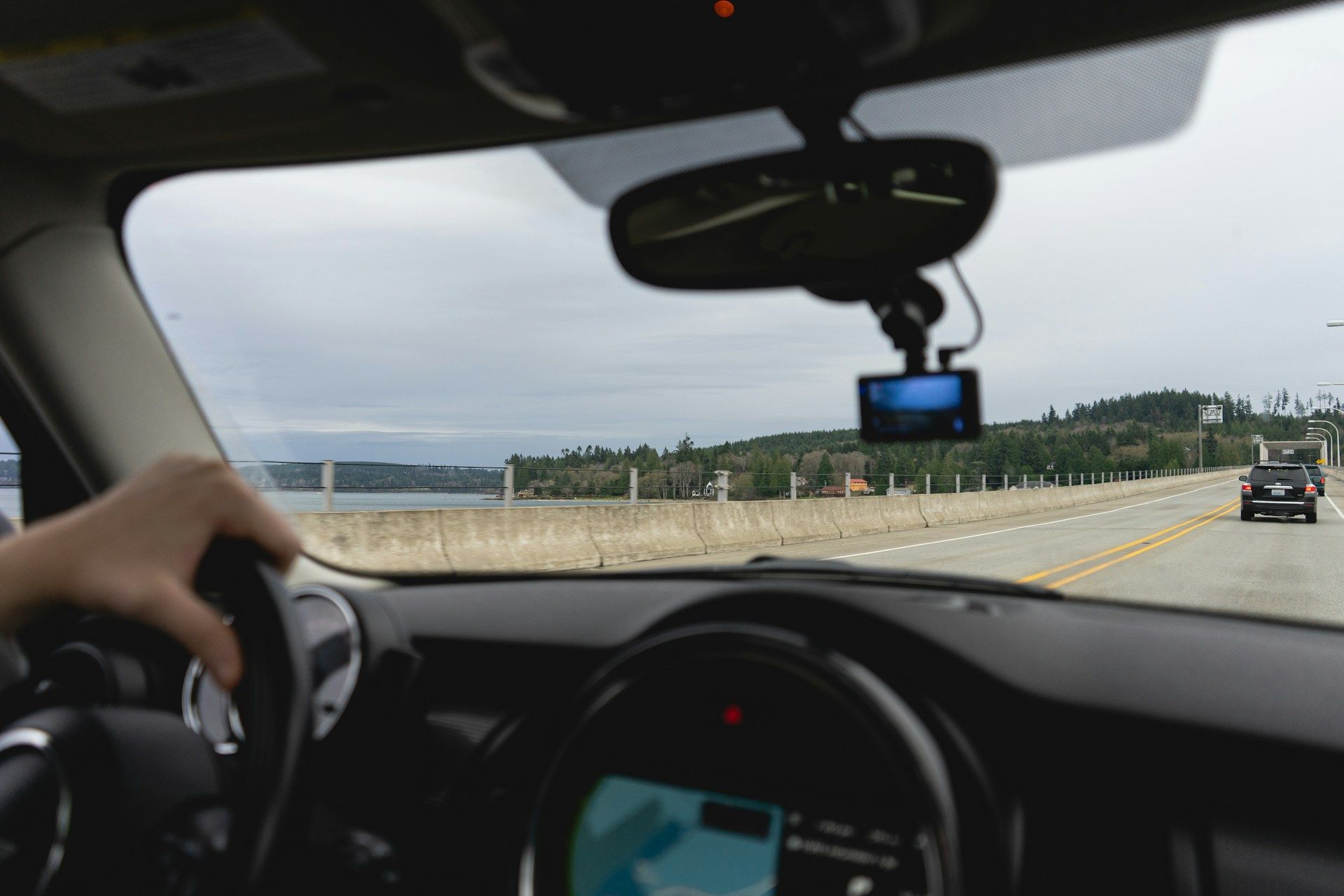 View from inside a car driving on a bridge. Cloudy sky and water visible. Hand on steering wheel.