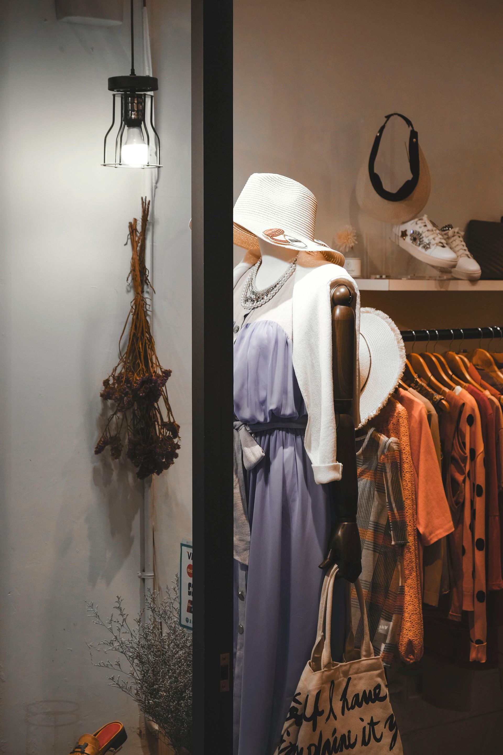Clothing store window display with mannequins wearing dresses and hats. Clothes hang on a rack.