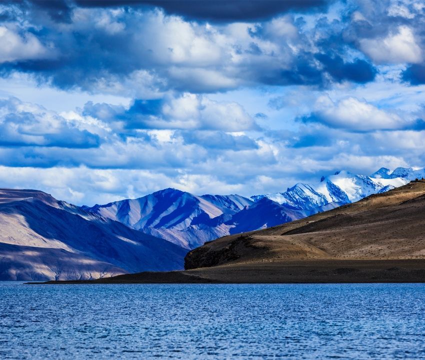 Scenic view of Pangong Tso Lake in Ladakh with tranquil blue water, barren hills, and snow-capped Himalayan peaks.