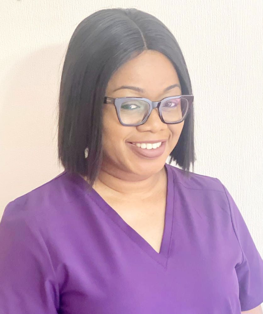 A woman wearing glasses and a purple scrub top is smiling.