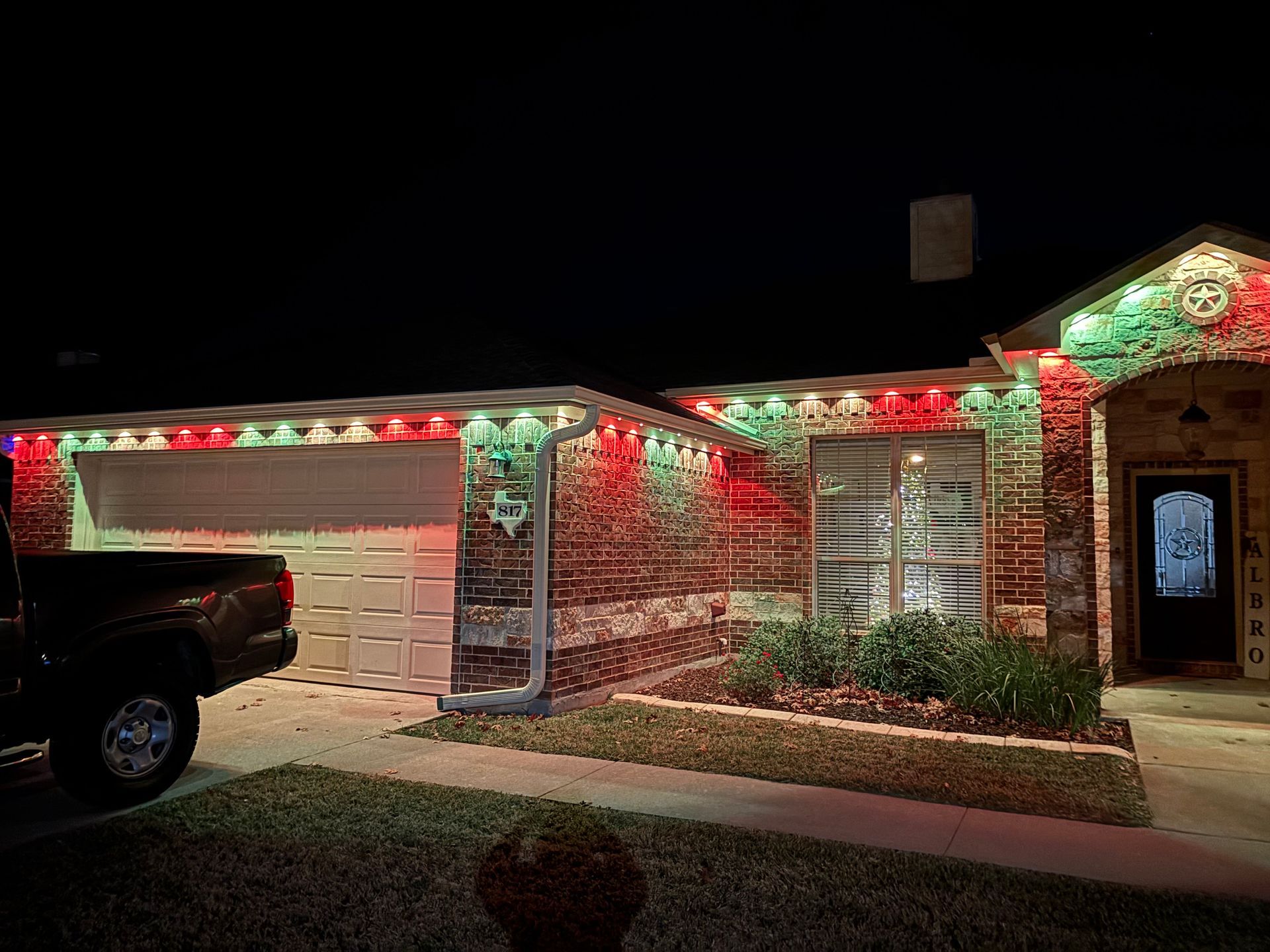 Single-story house at night decorated with red and green holiday lights, with a truck parked in the driveway.