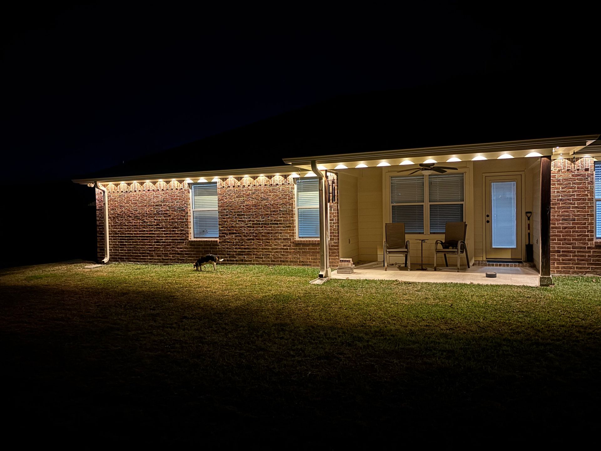 Brick house exterior at night, lit by warm porch lights, with a grassy yard in front.