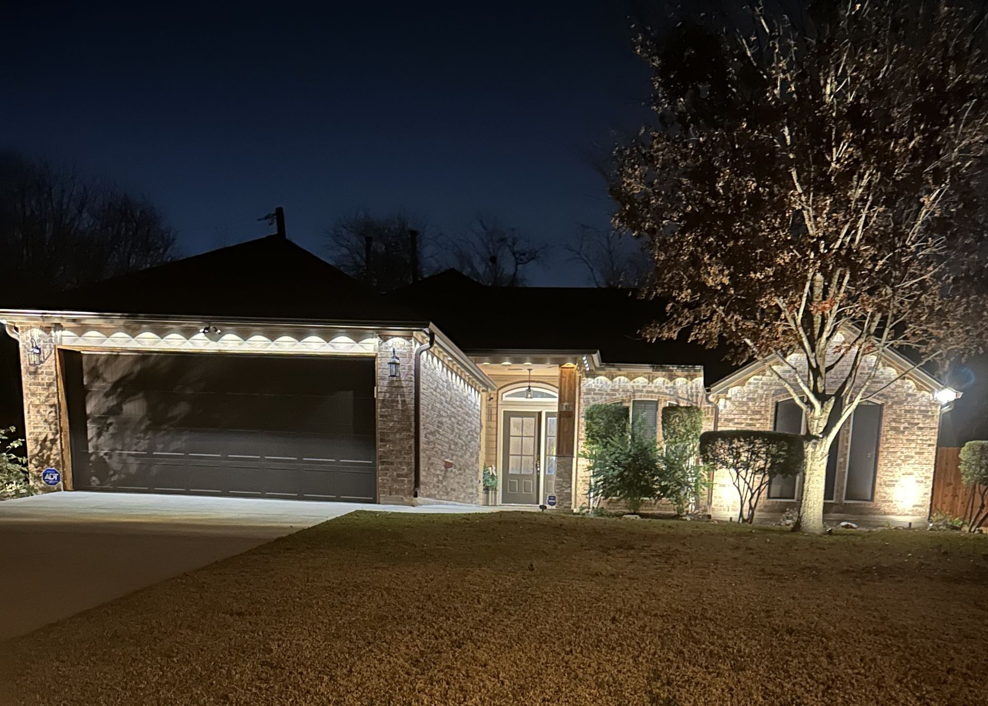 Night view of a brick house with lit garage, front porch, and a tree wrapped in white lights