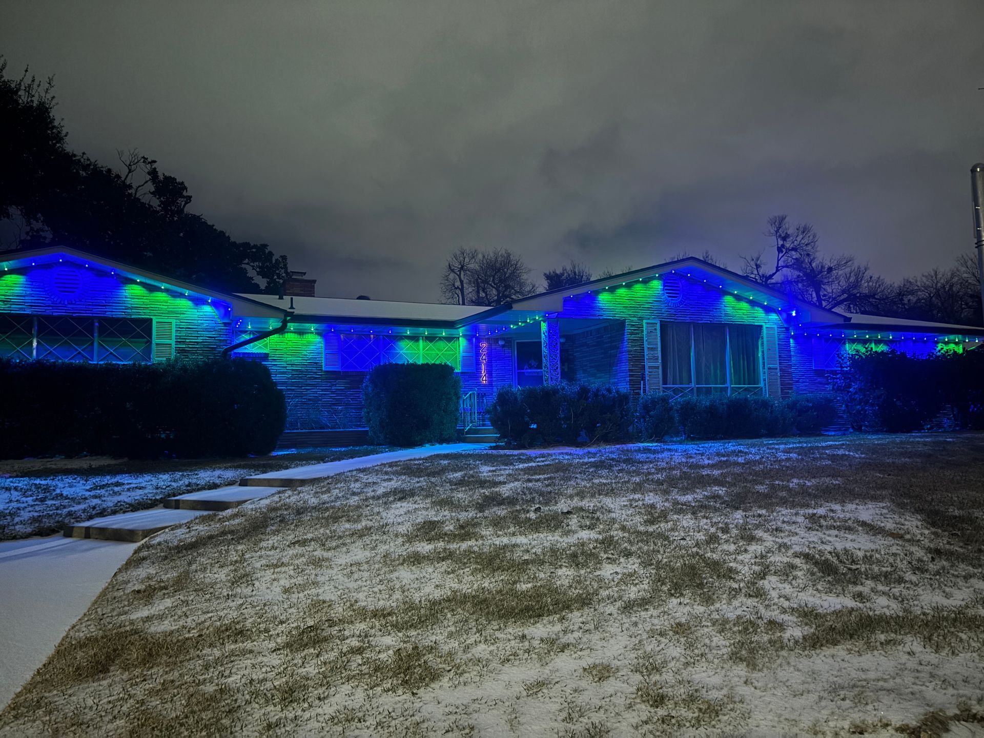 House lit with blue and green holiday lights at night, with a snowy front yard and cloudy sky.
