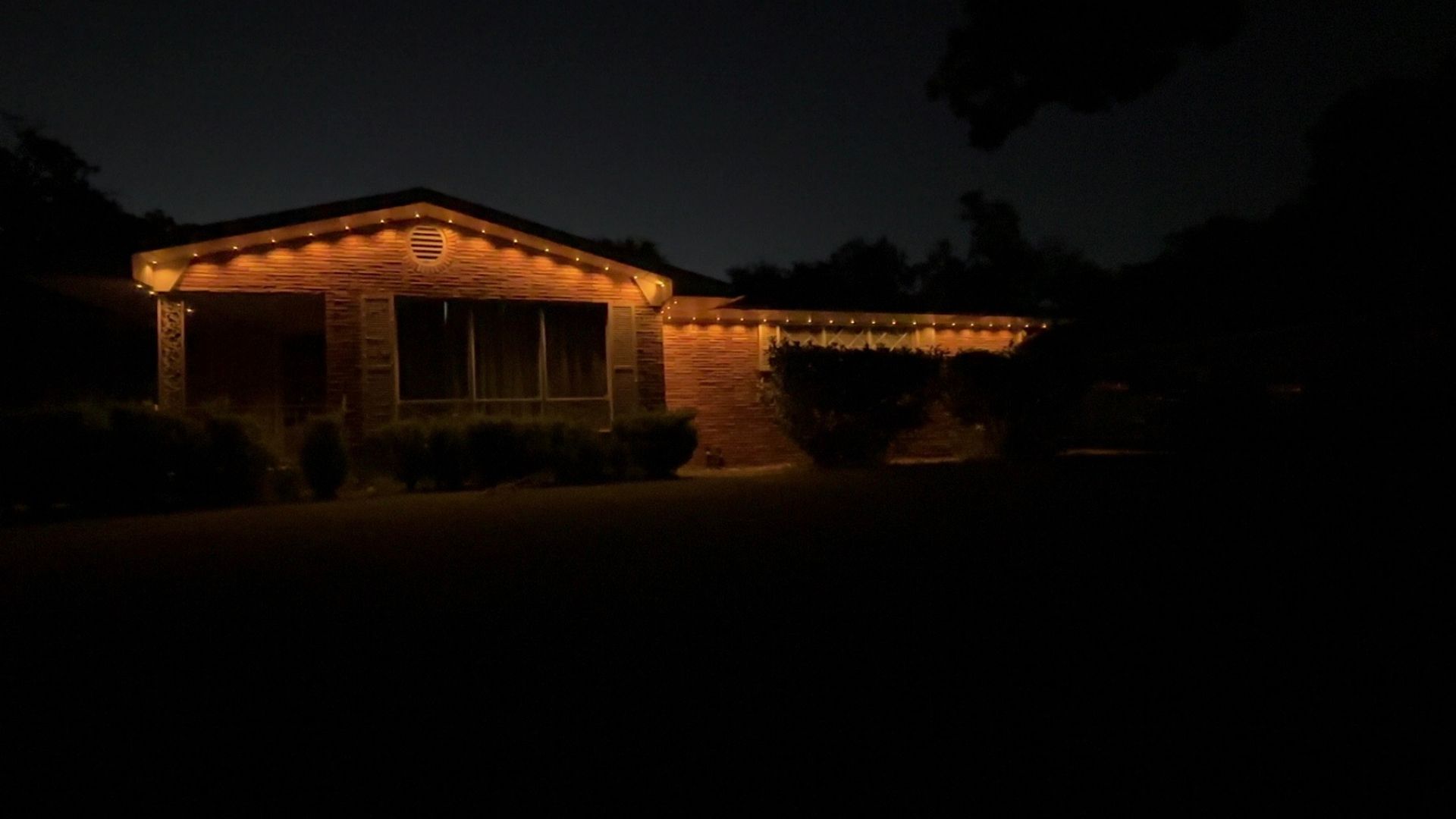 House outlined with warm white lights at night, with a dark yard and trees in the background