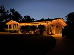House lit with warm orange string lights at night, surrounded by dark trees and a driveway.