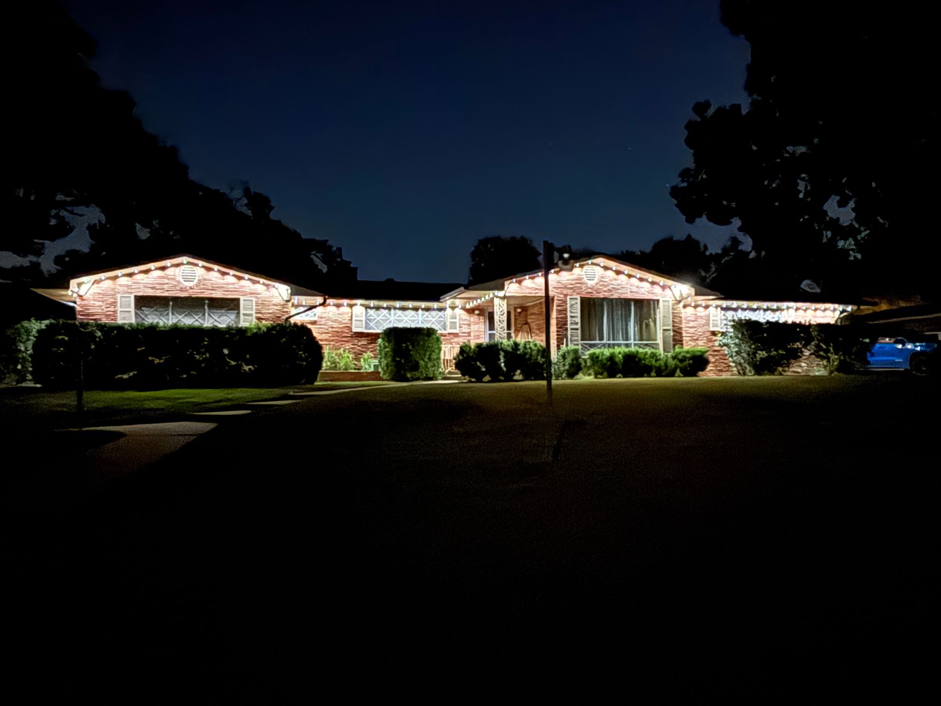 Single-story house lit with white holiday lights at night, surrounded by dark trees and lawn.